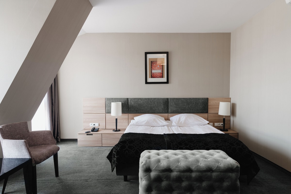 A hotel room with a bed, bedside lamps, a chair, a small table, and a framed picture on the wall.