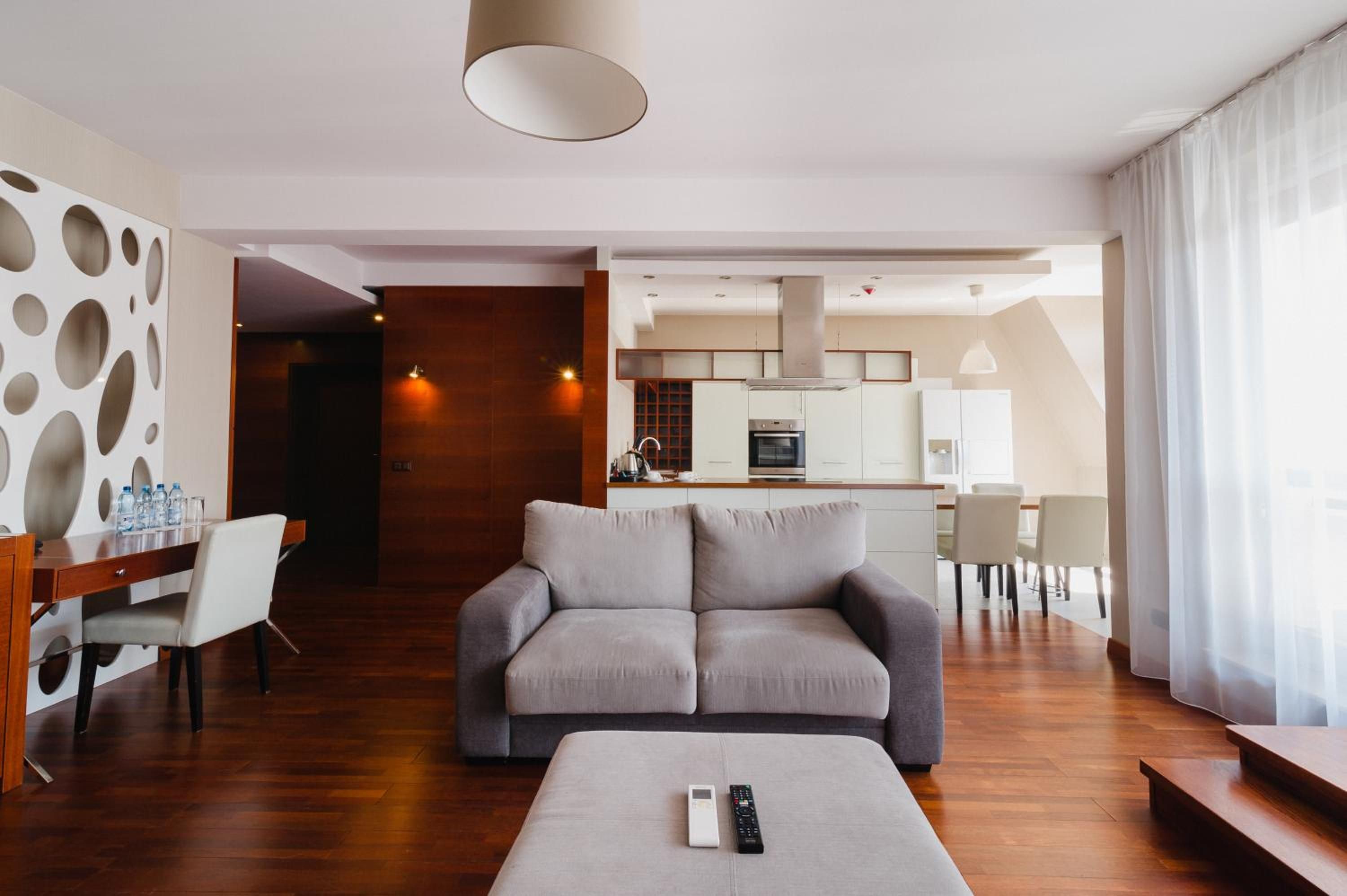 Ein modernes Wohnzimmer mit grauem Sofa, einem Esstisch mit Stühlen, einer Küche im Hintergrund und einem großen Fenster mit Vorhängen.