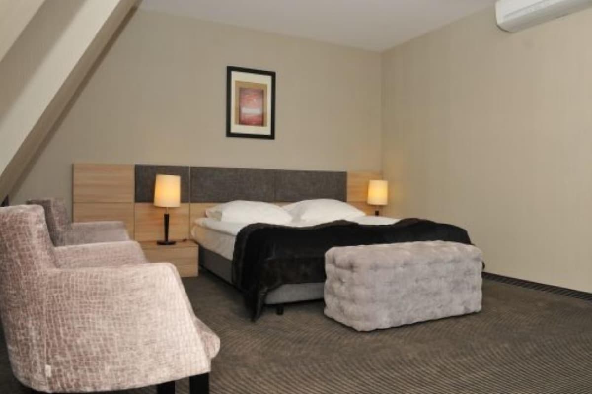 A hotel room with a bed, two bedside lamps, a headboard, a small ottoman, and a chair.