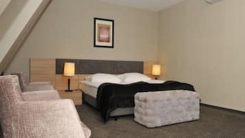 Quarto de hotel com cama, duas luminárias de cabeceira, encosto de cama, um banquinho pequeno e uma cadeira.