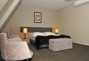 A hotel room with a bed, two bedside lamps, a headboard, a small ottoman, and a chair.