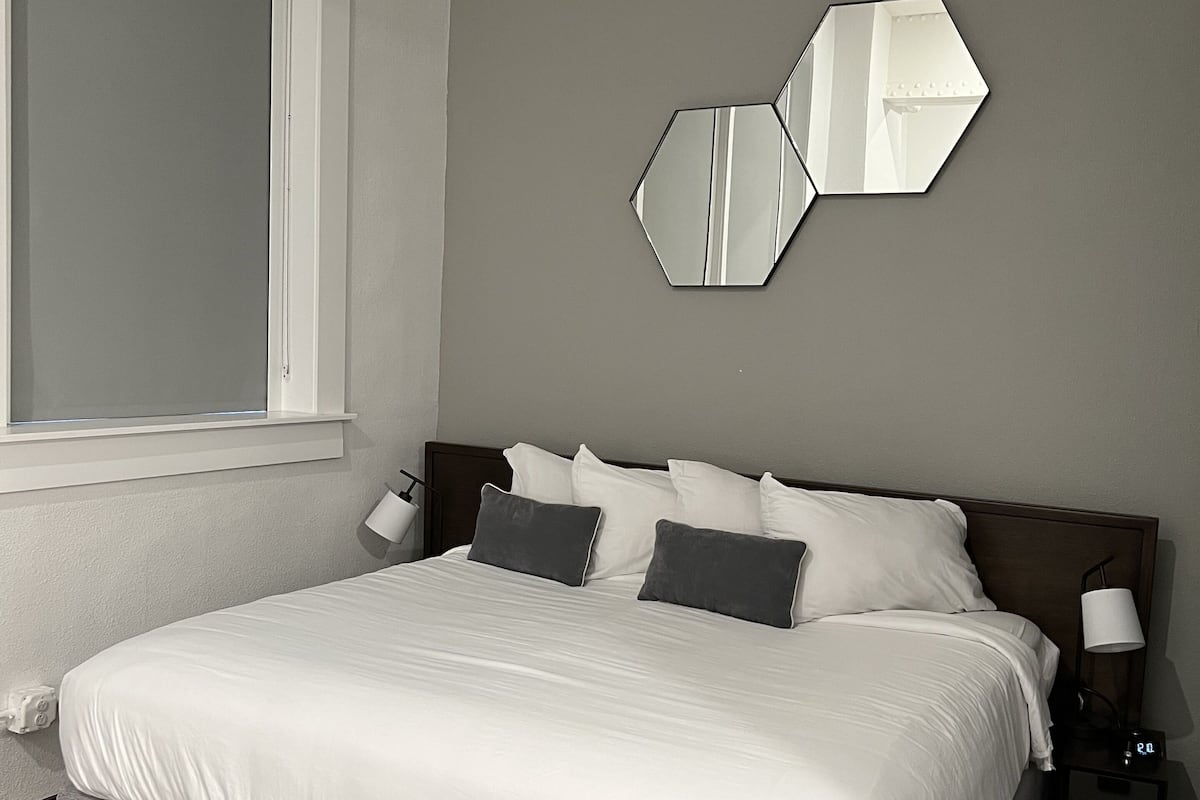 A neatly made bed with white bedding and two dark pillows, a wall-mounted hexagonal mirror, and a bedside table with a lamp and a digital clock.