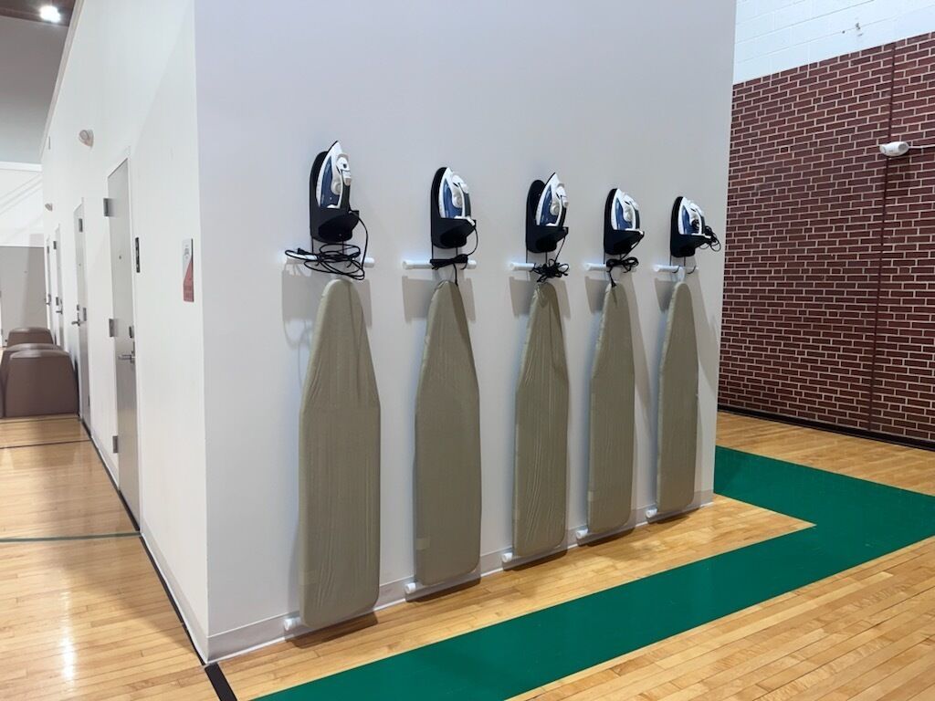 A row of wall-mounted ironing stations with a green carpeted floor and a brick wall in the background.