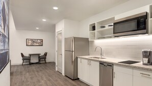 A modern kitchen with white cabinets, a stainless steel refrigerator, a microwave, and a dishwasher. There is a small dining area with a table and chairs.