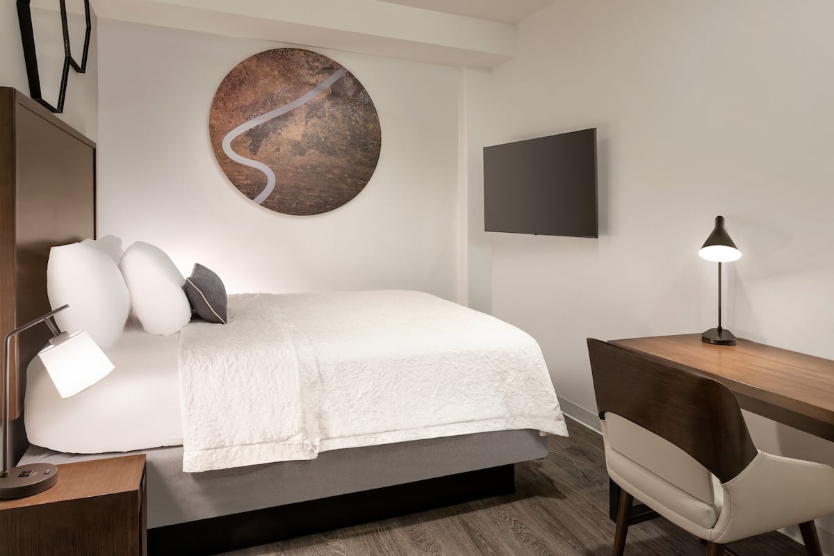A modern hotel room with a bed, a desk, a chair, a lamp, a TV, and a wall art piece.