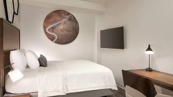 Una camera d'albergo moderna con un letto, una scrivania, una sedia, una lampada, una televisione e un'opera d'arte a parete.