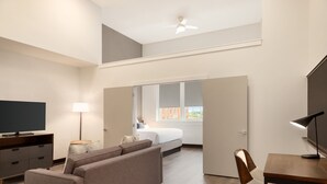 A modern hotel room with a sofa, a desk, a TV, a lamp, and a bed.