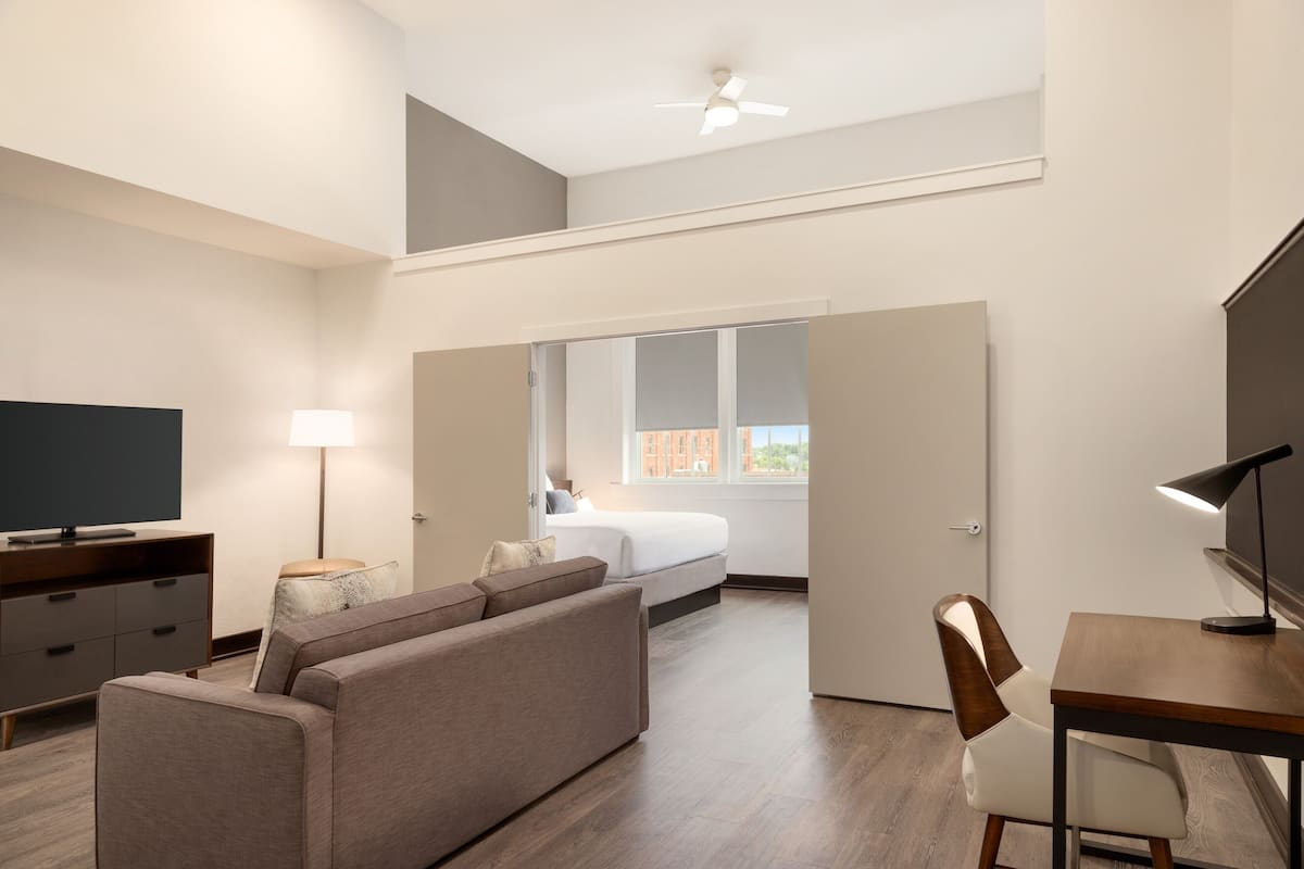 A modern hotel room with a sofa, a desk, a TV, a lamp, and a bed.