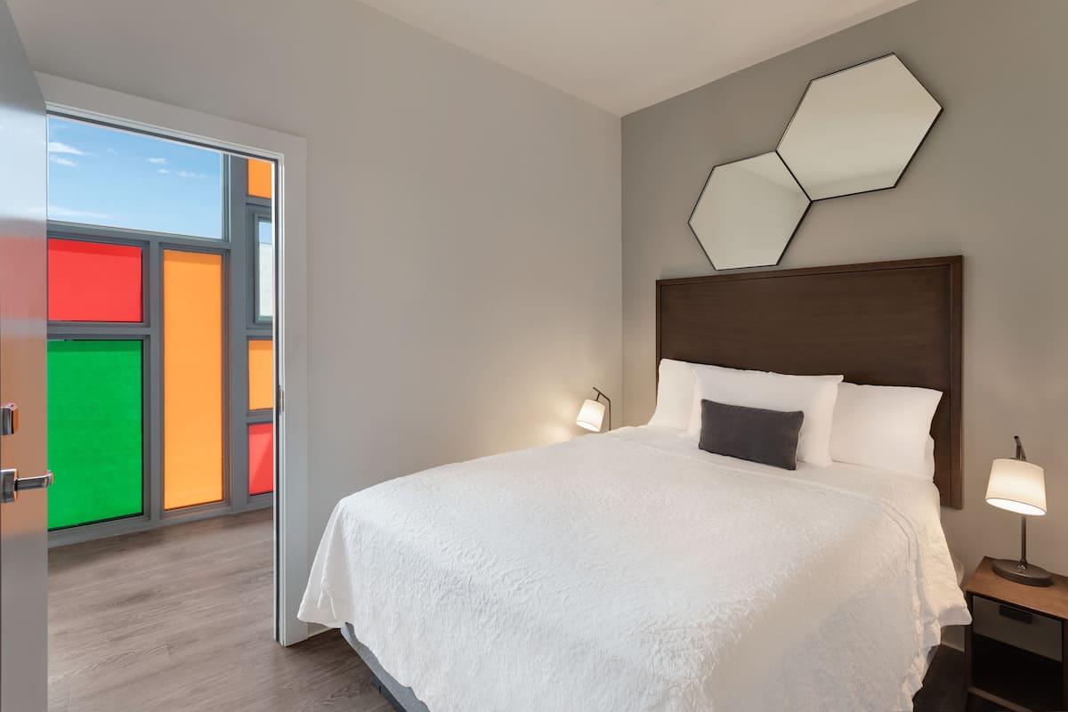 A modern hotel room with a large bed, a nightstand, a wall-mounted mirror, and a view through a door with colorful panels.