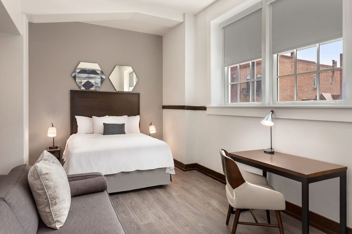 A hotel room with a bed, a desk, a chair, a sofa, and a window with a view of buildings.