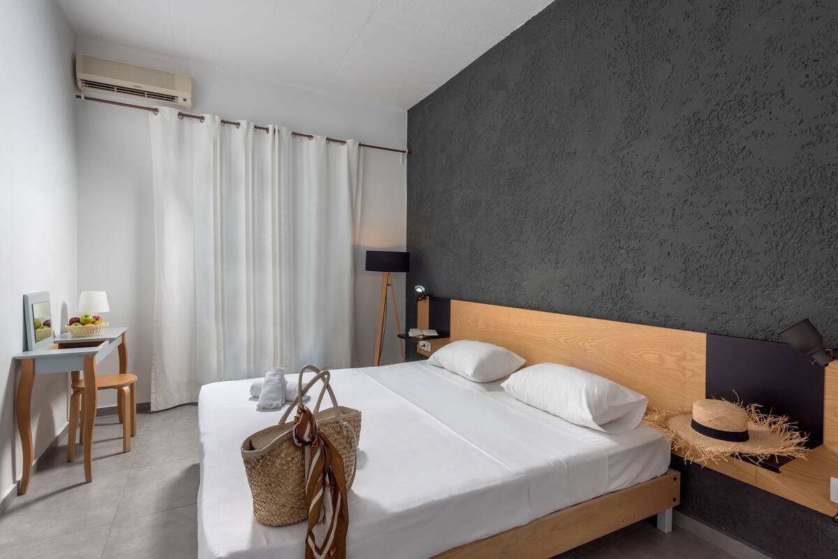 A modern bedroom with a wooden bed, white bedding, a desk with a chair, and a small table with fruit and a lamp.