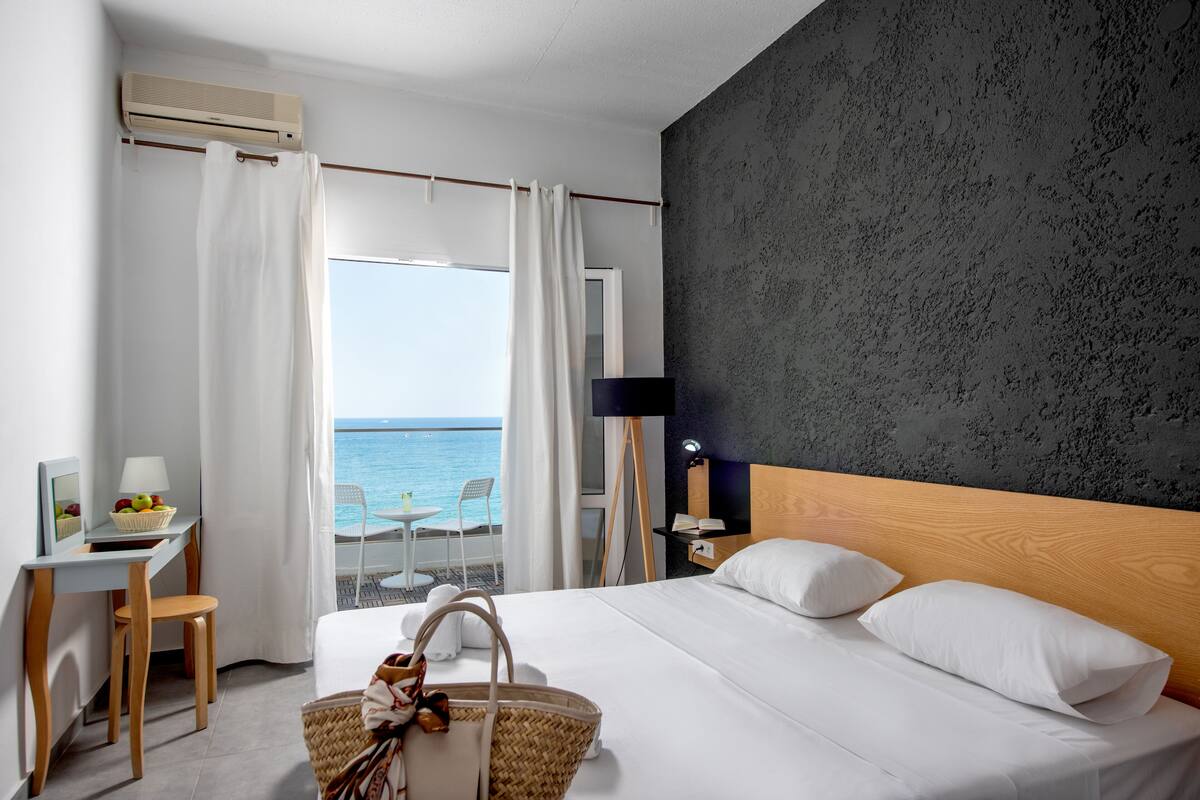 A hotel room with a bed, a desk, a chair, a lamp, and a view of the sea.