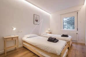 Un dormitorio con dos camas, mesitas de noche, un radiador y una ventana con vistas a árboles.