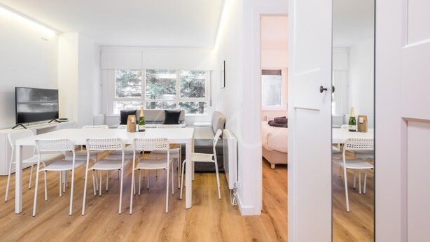 A modern, well-lit living room with a large white table, white chairs, a sofa, and a TV. - Saboredo (Baqueira)