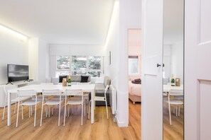 Una sala de estar moderna y bien iluminada con una mesa blanca grande, sillas blancas, un sofá y un televisor.
