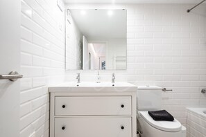 Un baño moderno con paredes de azulejos blancos, un lavabo blanco con dos grifos, un espejo sobre el lavabo y un inodoro con una toalla.