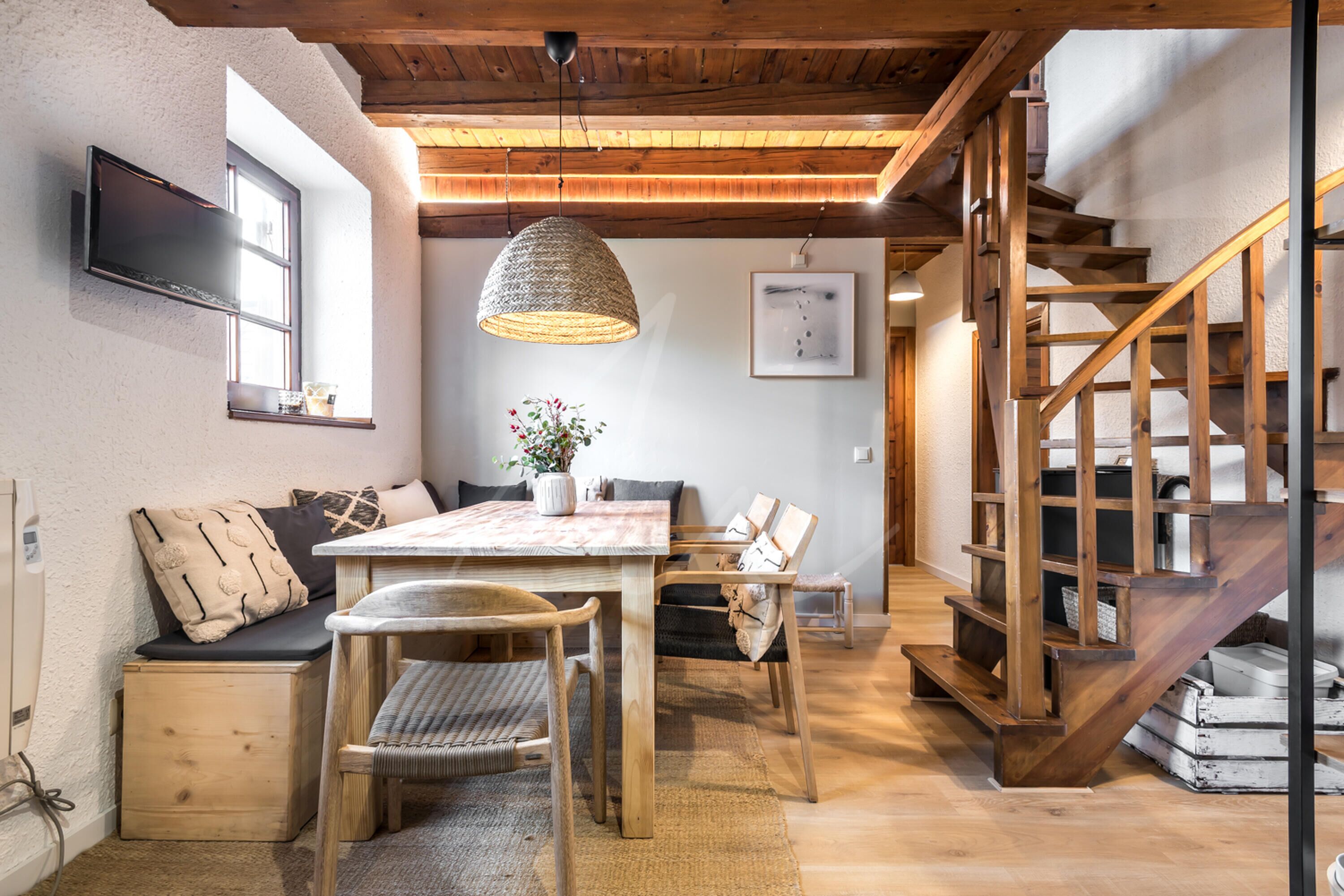 A cozy dining area with a wooden table and chairs, a woven pendant light, and a staircase with wooden railings.