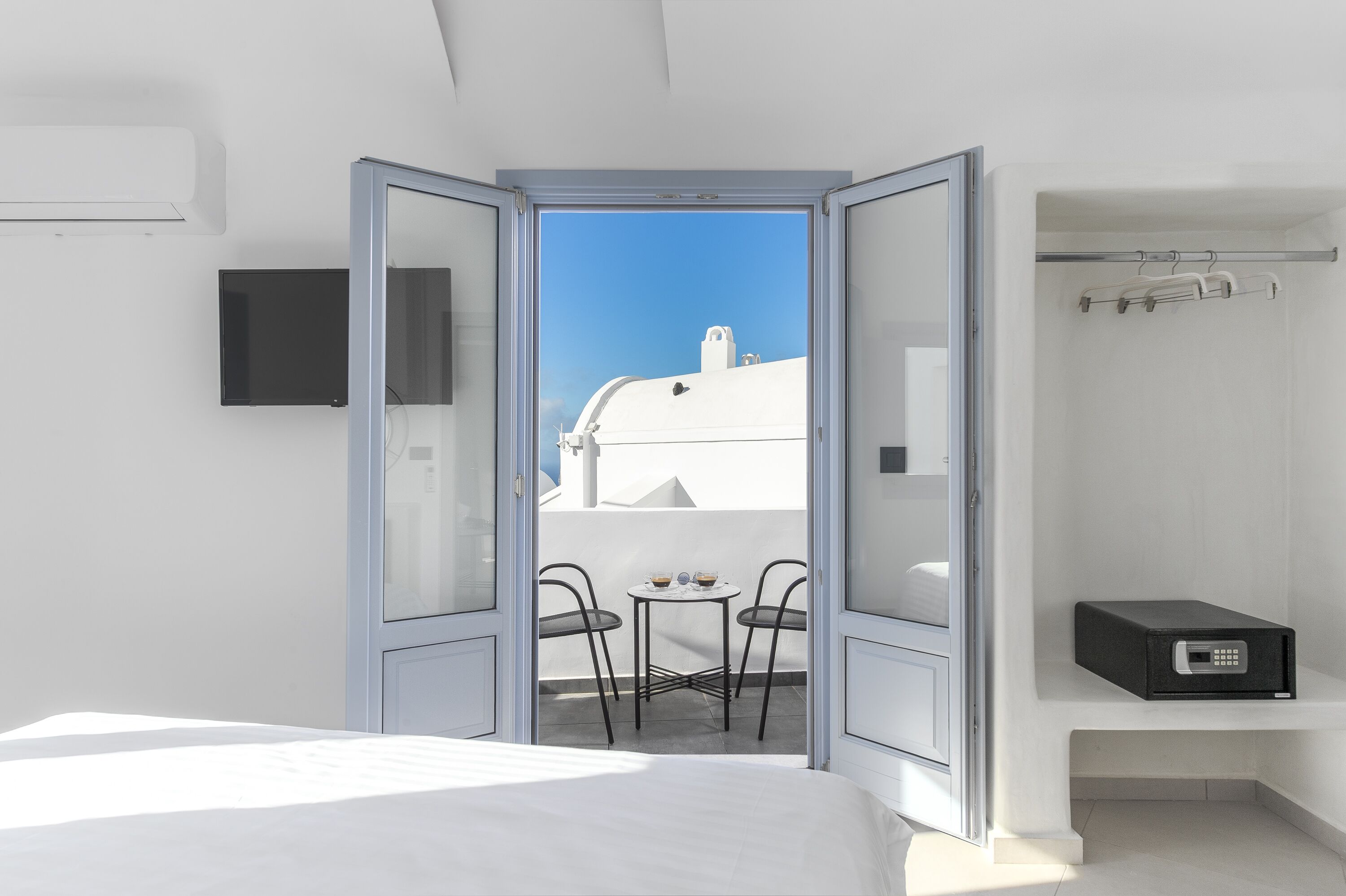 Habitación de hotel con cama, una mesita con sillas, televisión y vistas al cielo y edificios a través de una puerta abierta.