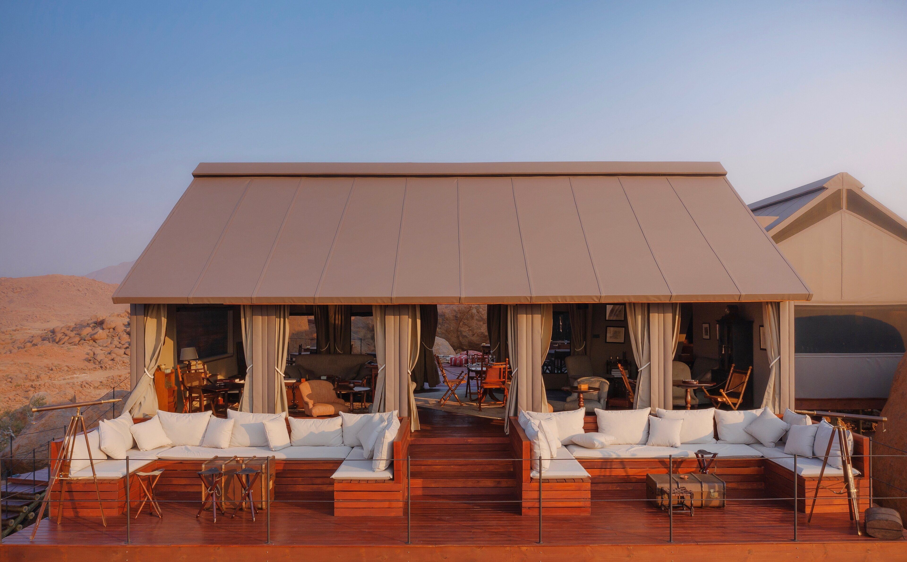 Un amplio espacio al aire libre con varios sofás y sillones, una terraza de madera y una gran estructura tipo tienda que proporciona sombra.