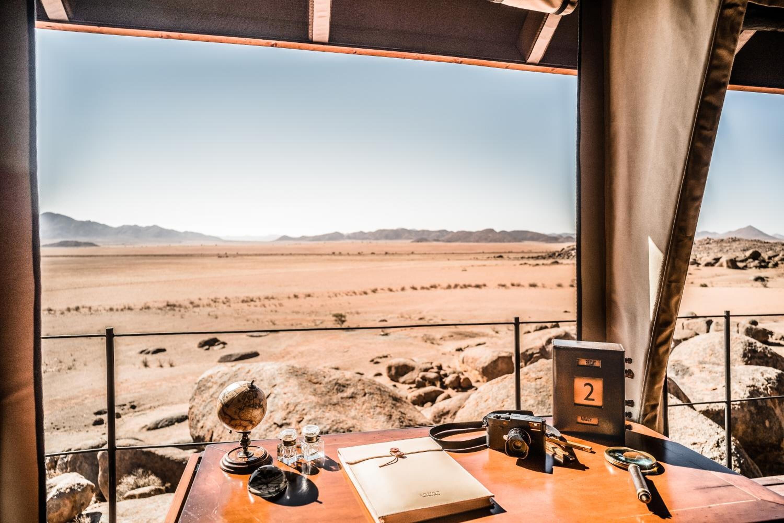 Une vue sur le désert, prise depuis une fenêtre donnant sur un bureau où se trouvent un globe terrestre, un carnet, un appareil photo et un panneau portant le chiffre 2.