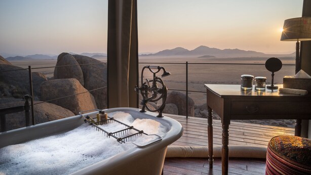 A bathtub with a view of a desert landscape, a table with jars, and a lamp. - Zannier Sonop (Sesriem)