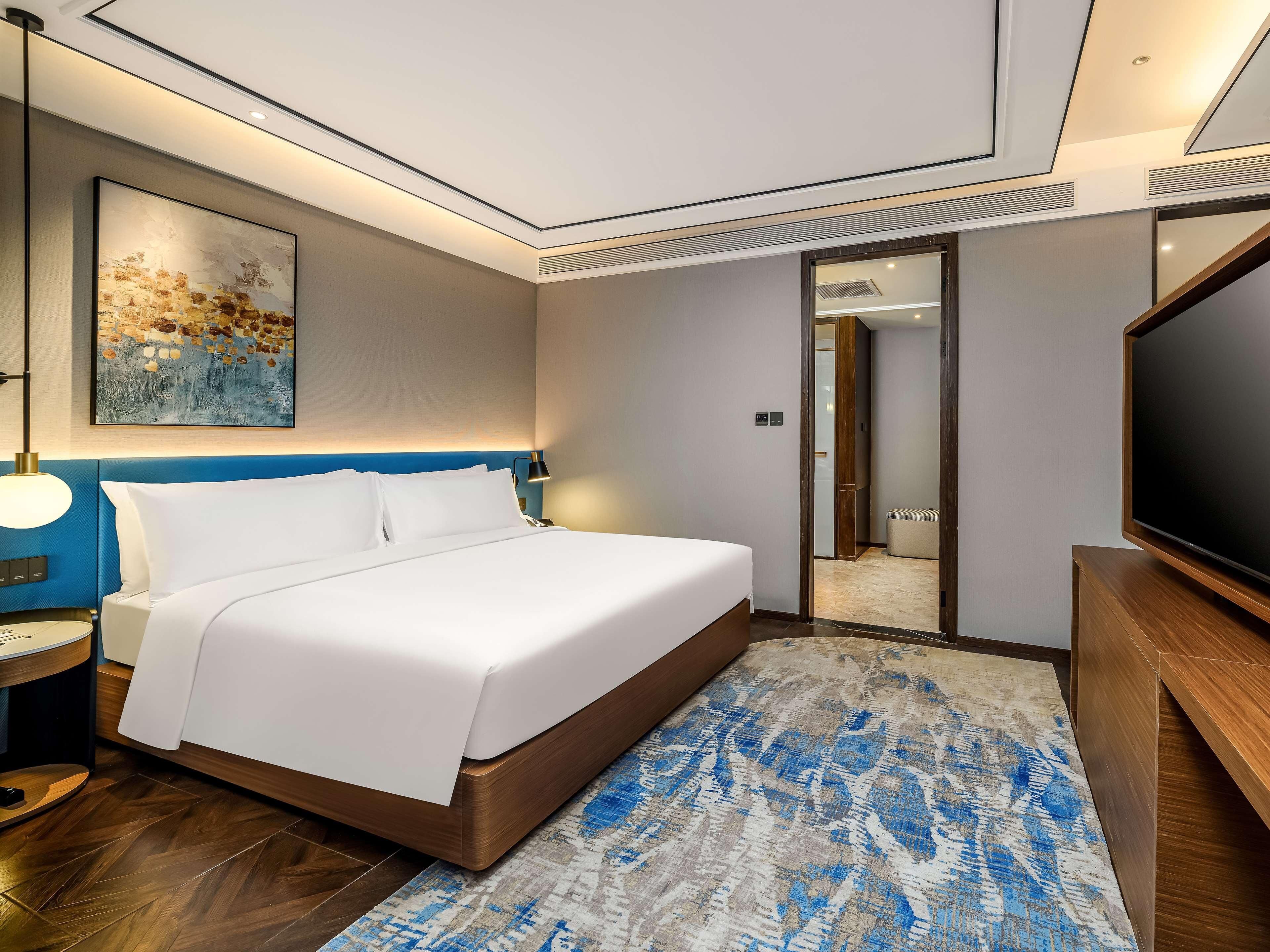A modern hotel room with a large bed, a flat-screen TV, a desk with a lamp, and a painting on the wall.