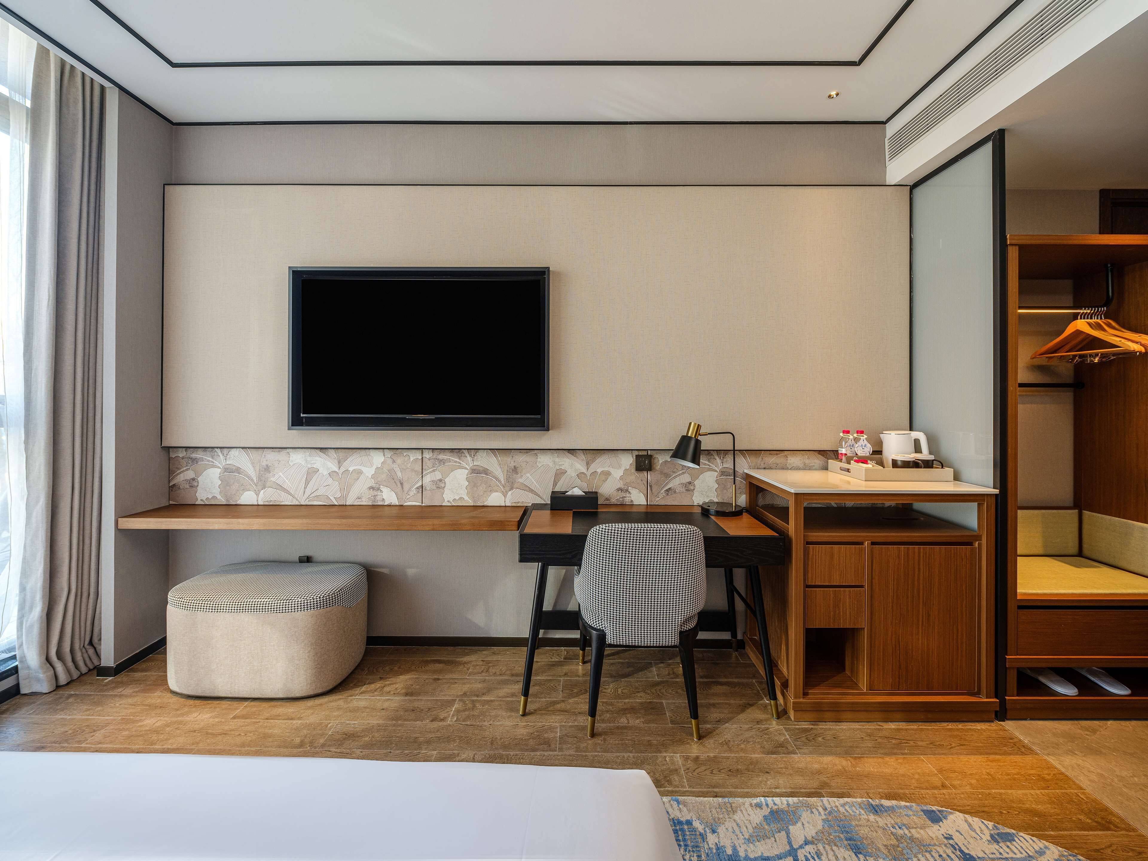 A modern hotel room with a flat-screen TV mounted on the wall, a wooden desk, a chair, a small ottoman, and a walk-in closet with a hanging rack.