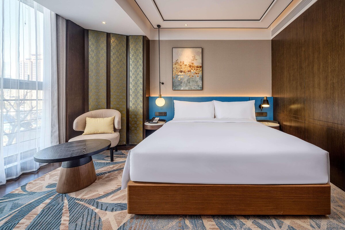 A hotel room with a large bed, a chair, a small table, and a view of the city.