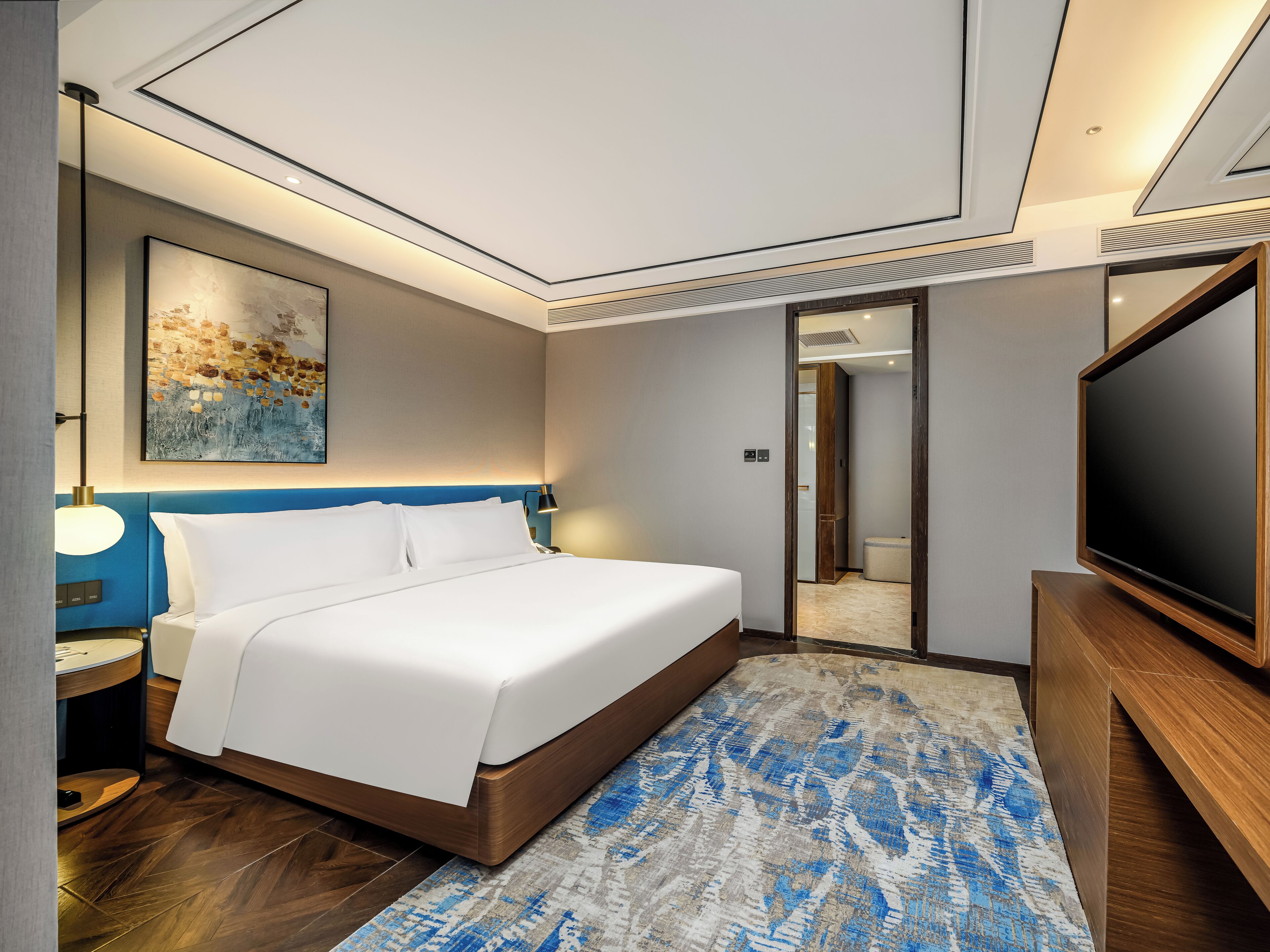 Une chambre d’hôtel moderne dotée d’un grand lit, d’un téléviseur à écran plat, d’un tableau au mur et d’une salle de bain visible à travers une porte en verre.
