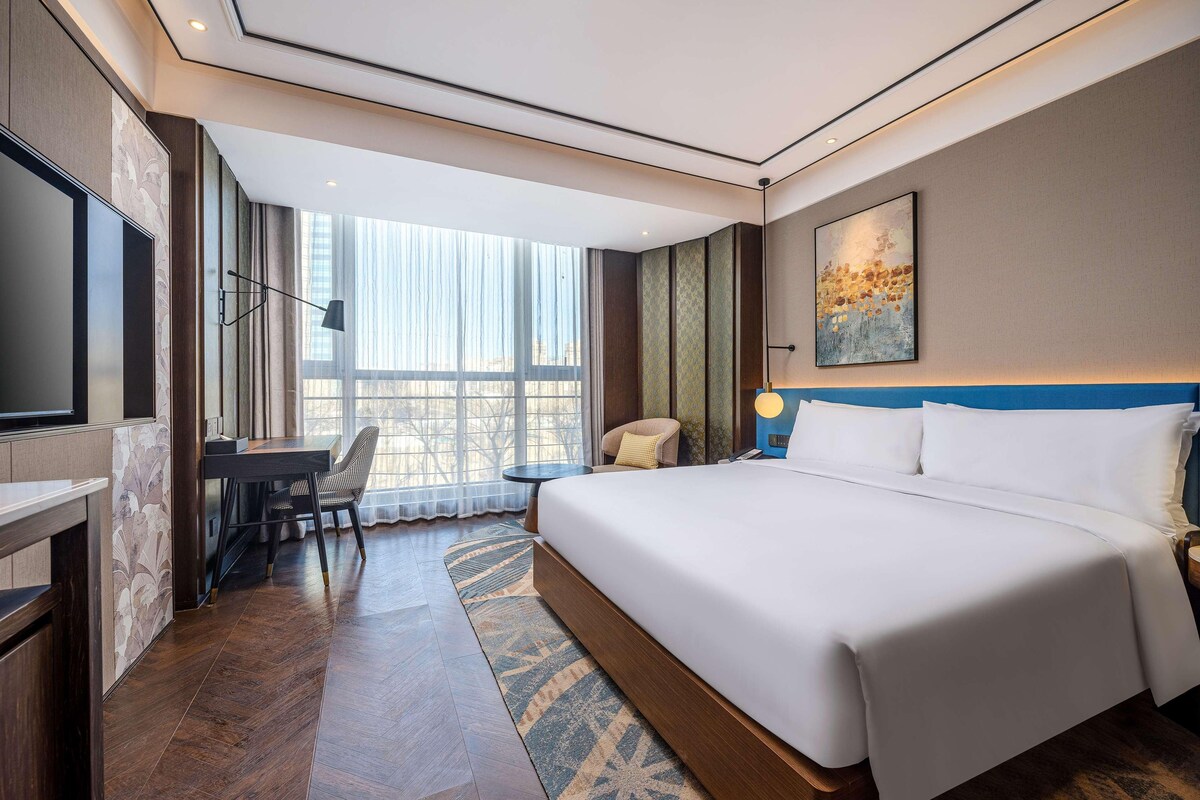 A hotel room with a large bed, a desk, a chair, a TV, and a large window.