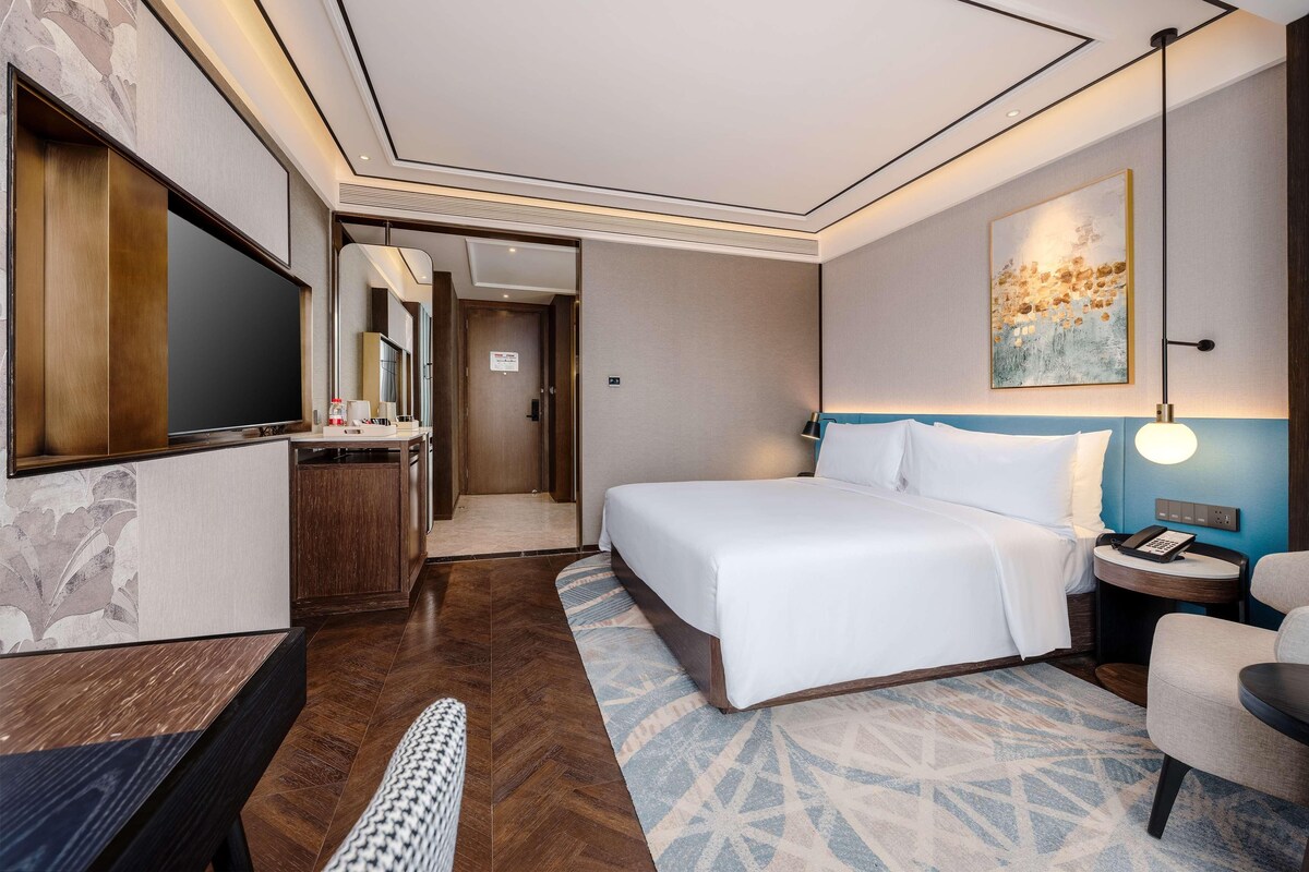 A hotel room with a large bed, a desk with a flat-screen TV, a bedside table with a lamp, and a wall-mounted artwork.