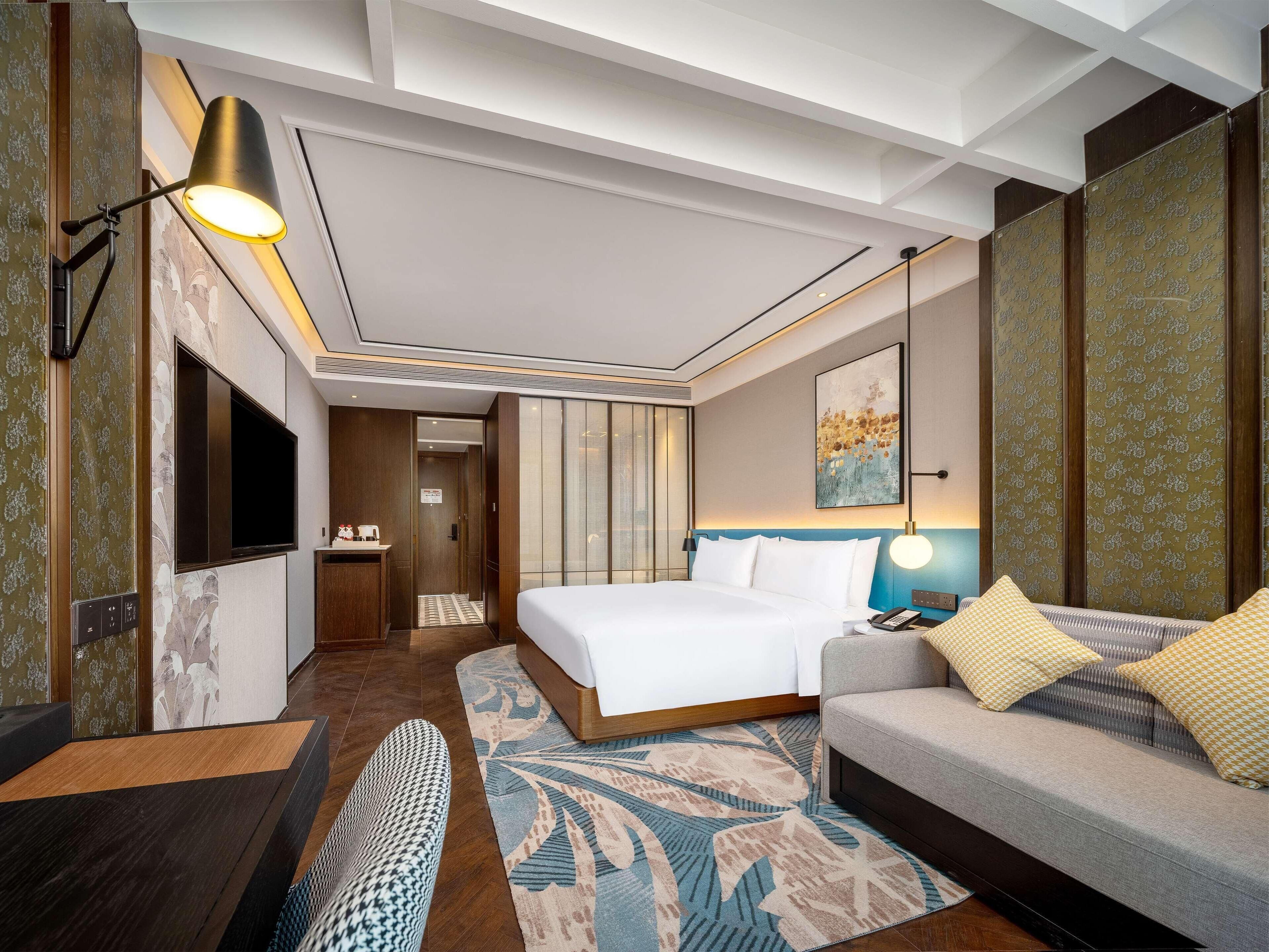 A modern hotel room with a bed, sofa, TV, and a desk with a lamp.