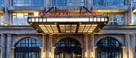 The entrance of the Hilton Garden Inn with a large marquee sign, arched windows, and a grand entrance with steps leading up to the doors.