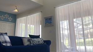 A spacious living room with a blue sofa, a patterned throw pillow, and a dark striped rug. There are large windows with sheer curtains, a ceiling fan, and a wall-mounted light fixture. - 7BR Family & Spacious Group Retreat!  Sleeps 14! Private Pool! 15 Min to Disney! (Kissimmee)