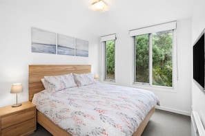 A bedroom with a wooden bed, two bedside tables, a night lamp, and a large window with a view of greenery. - Point Grey Apartment 7 (Lorne)