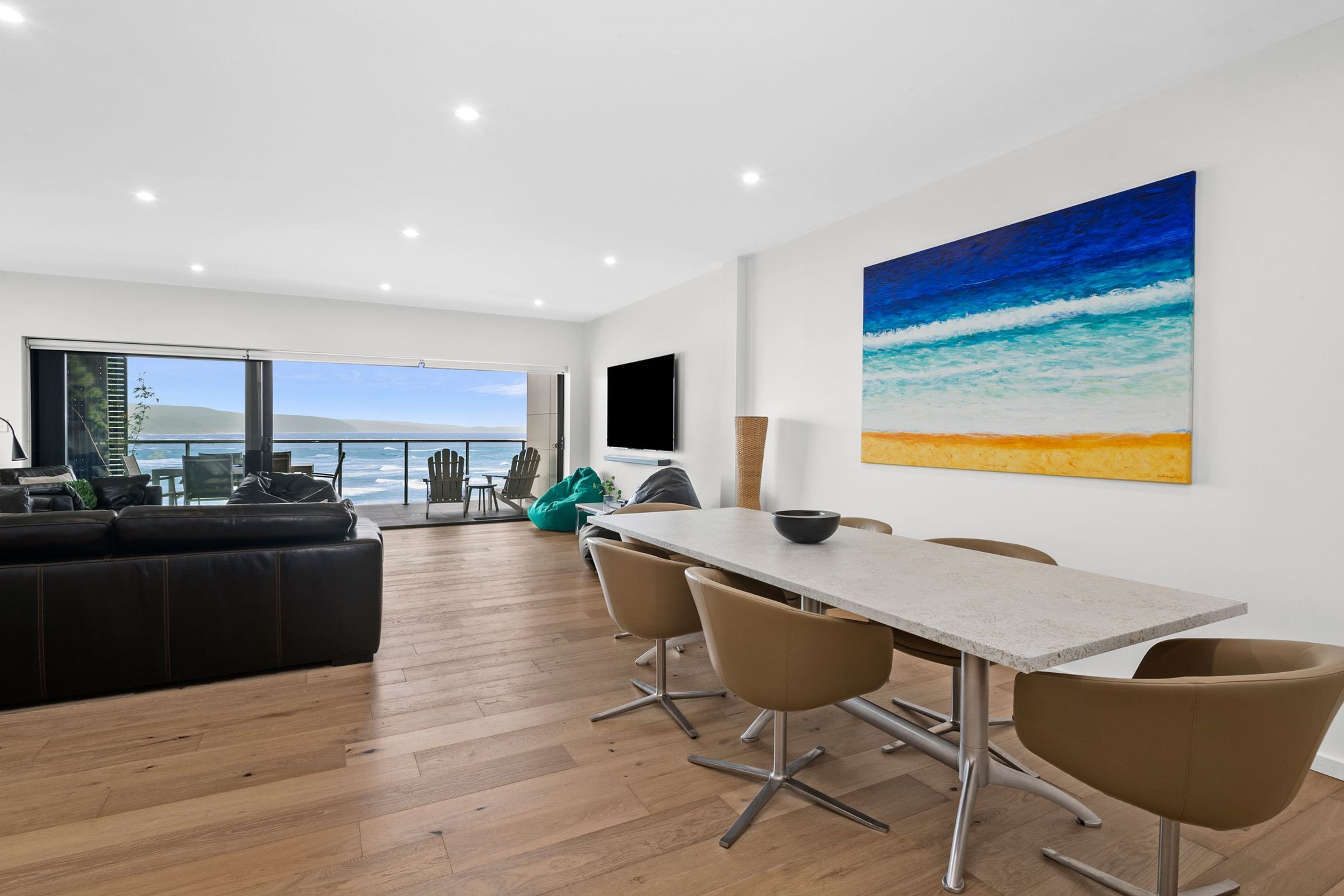A modern living room with a large window overlooking a body of water, a dining area with a table and chairs, and a large painting of a beach scene.
