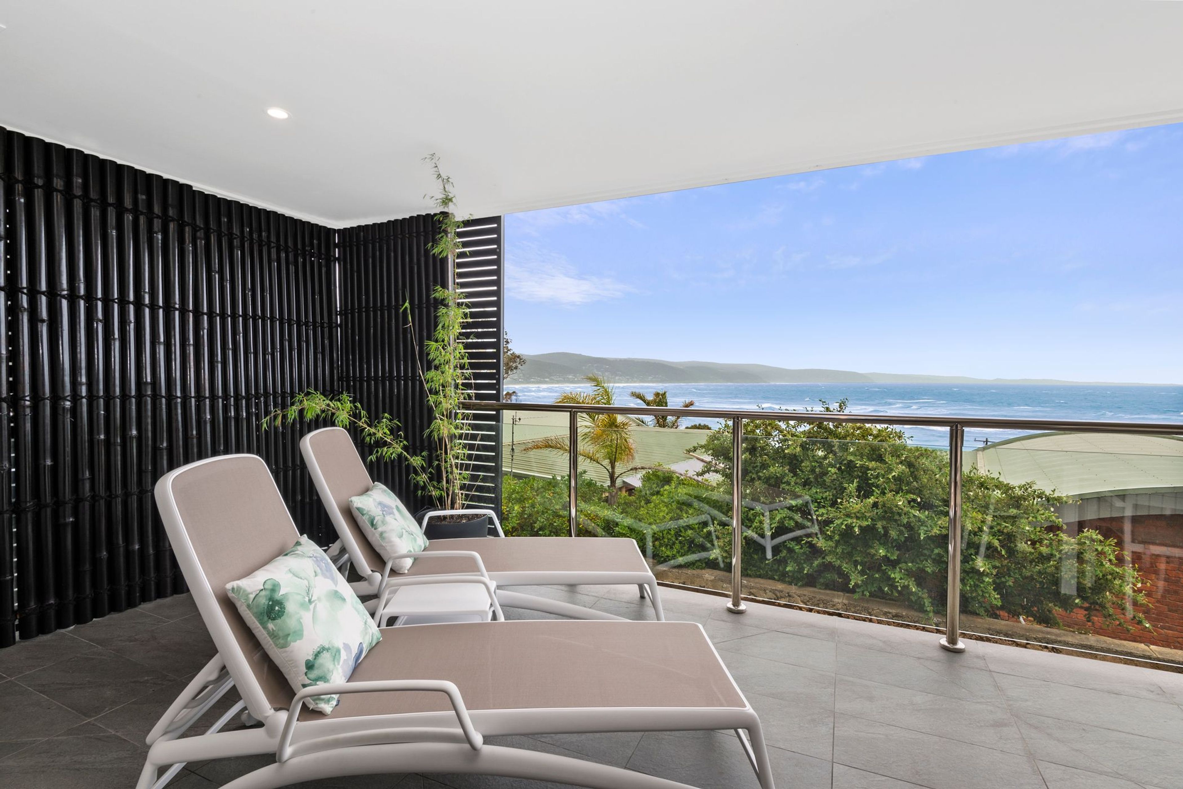 Ein Balkon mit drei Liegestühlen, einer schwarzen Bambuswand und Blick aufs Meer.