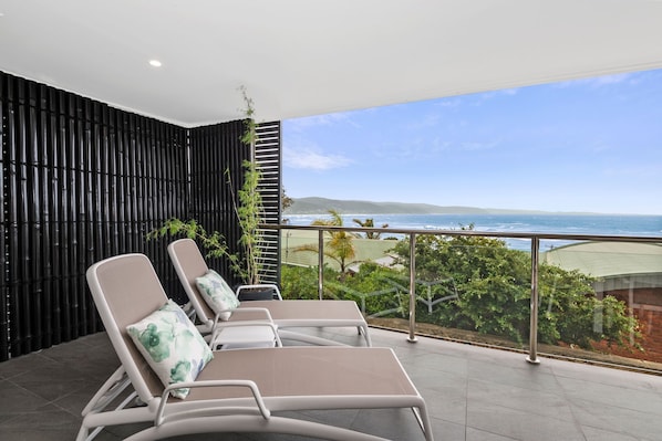 Ein Balkon mit drei Liegestühlen, einer schwarzen Bambuswand und Blick aufs Meer.