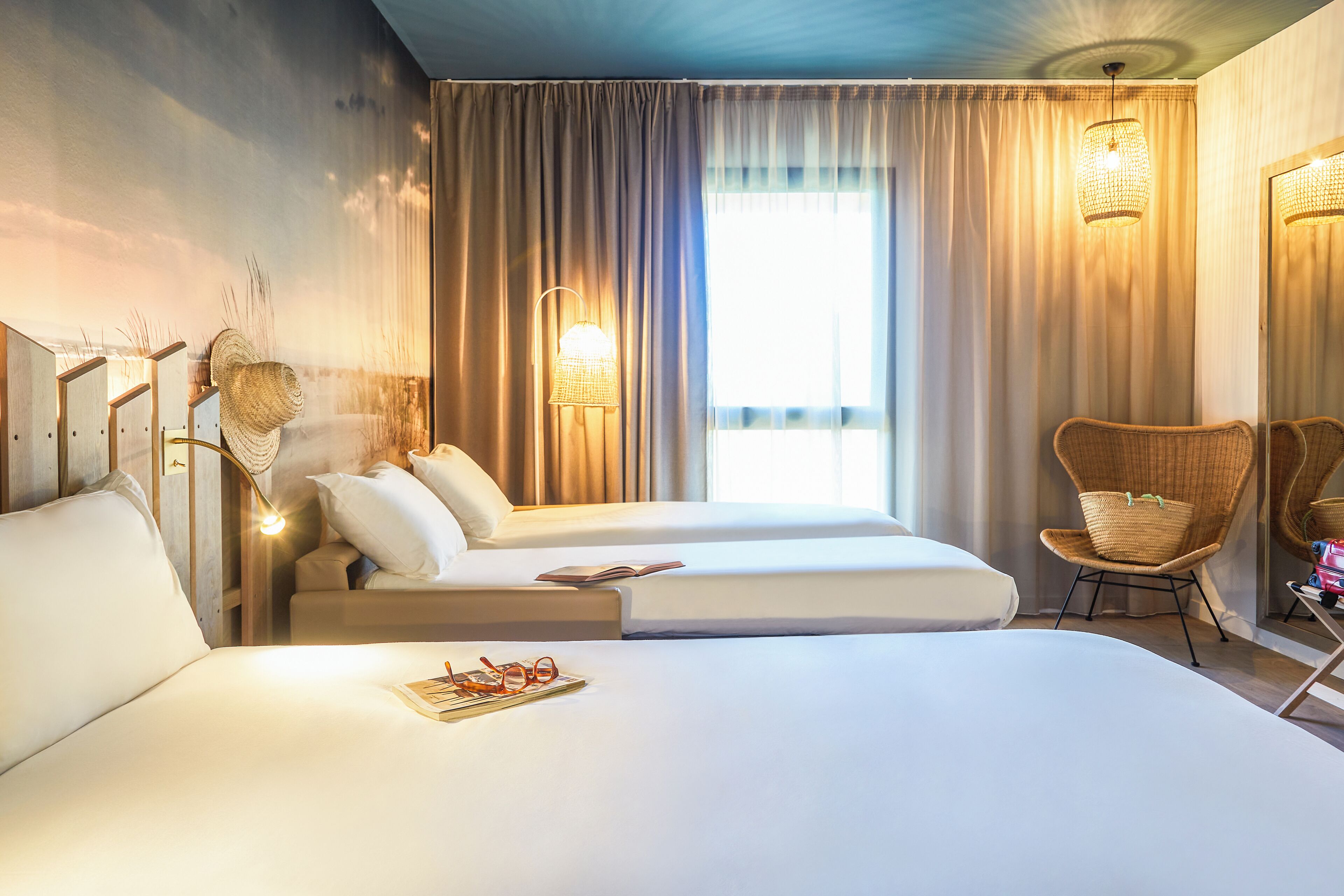 Habitación de hotel con dos camas, un ventanal con cortinas, una silla de mimbre y una lámpara.