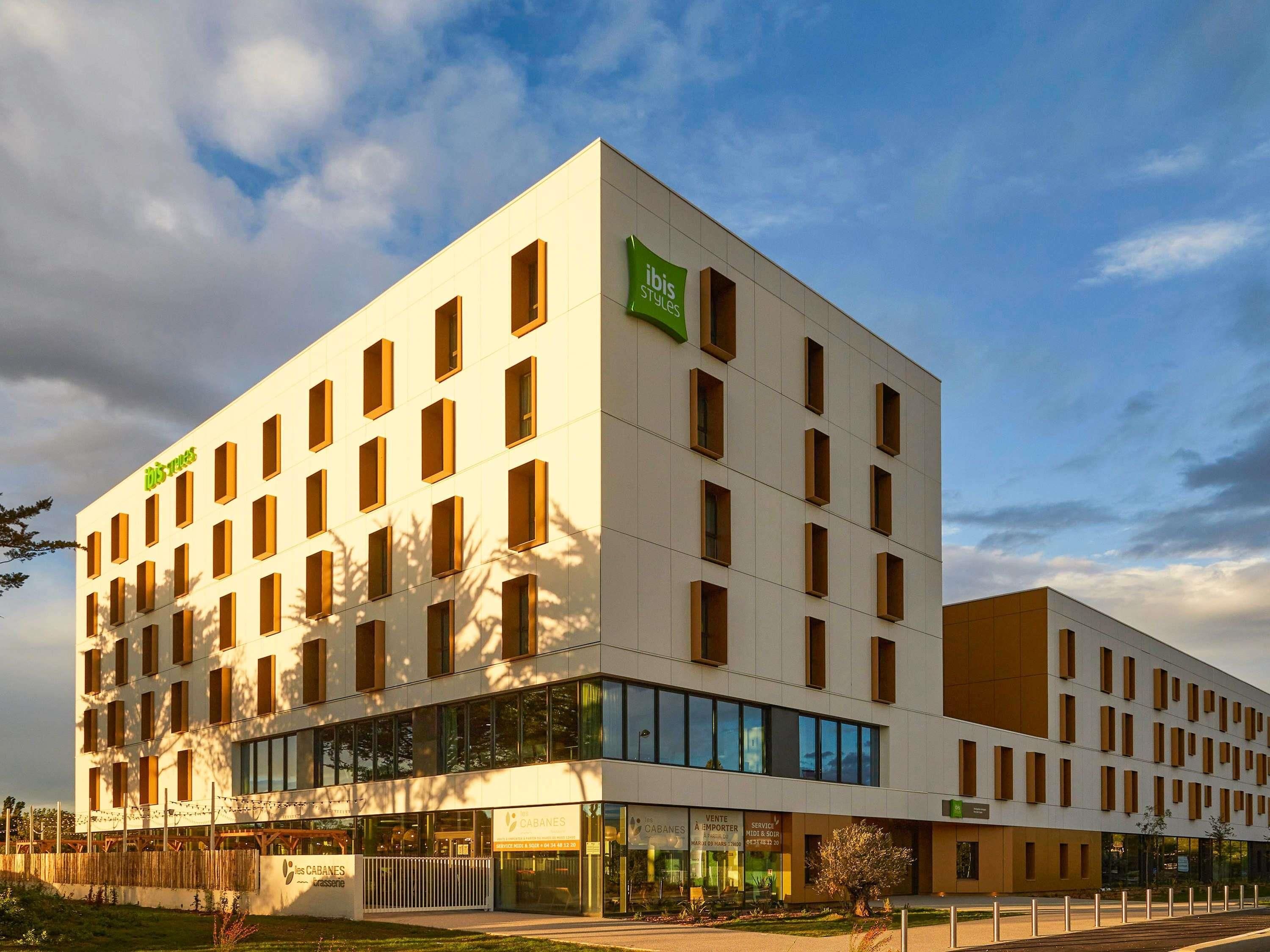 A modern hotel building with multiple floors, large windows, and a prominent green sign for ibis styles.