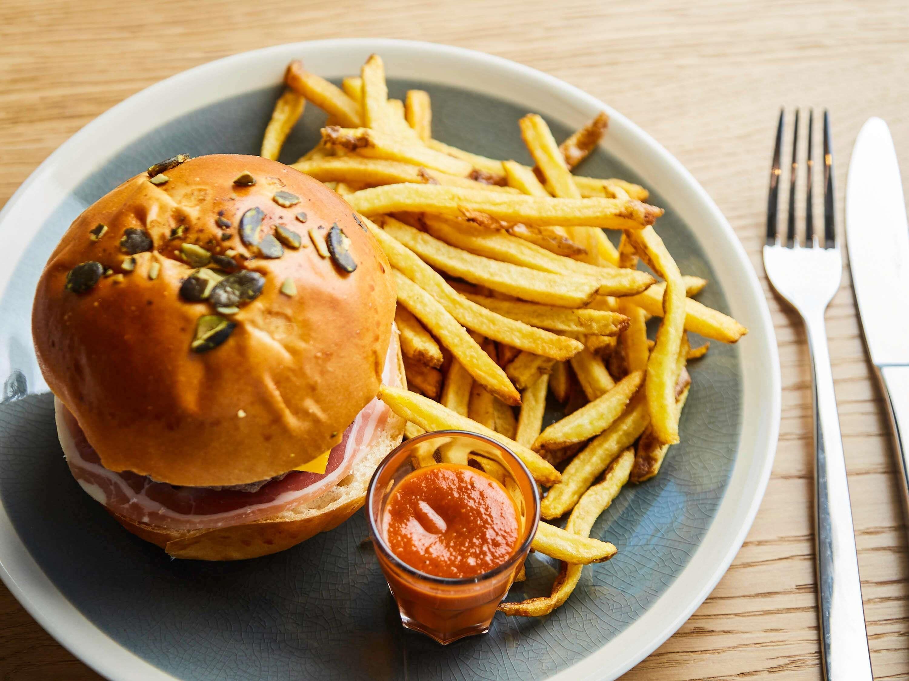 A plate with a hamburger and fries, a small glass of ketchup, and cutlery on a wooden table.