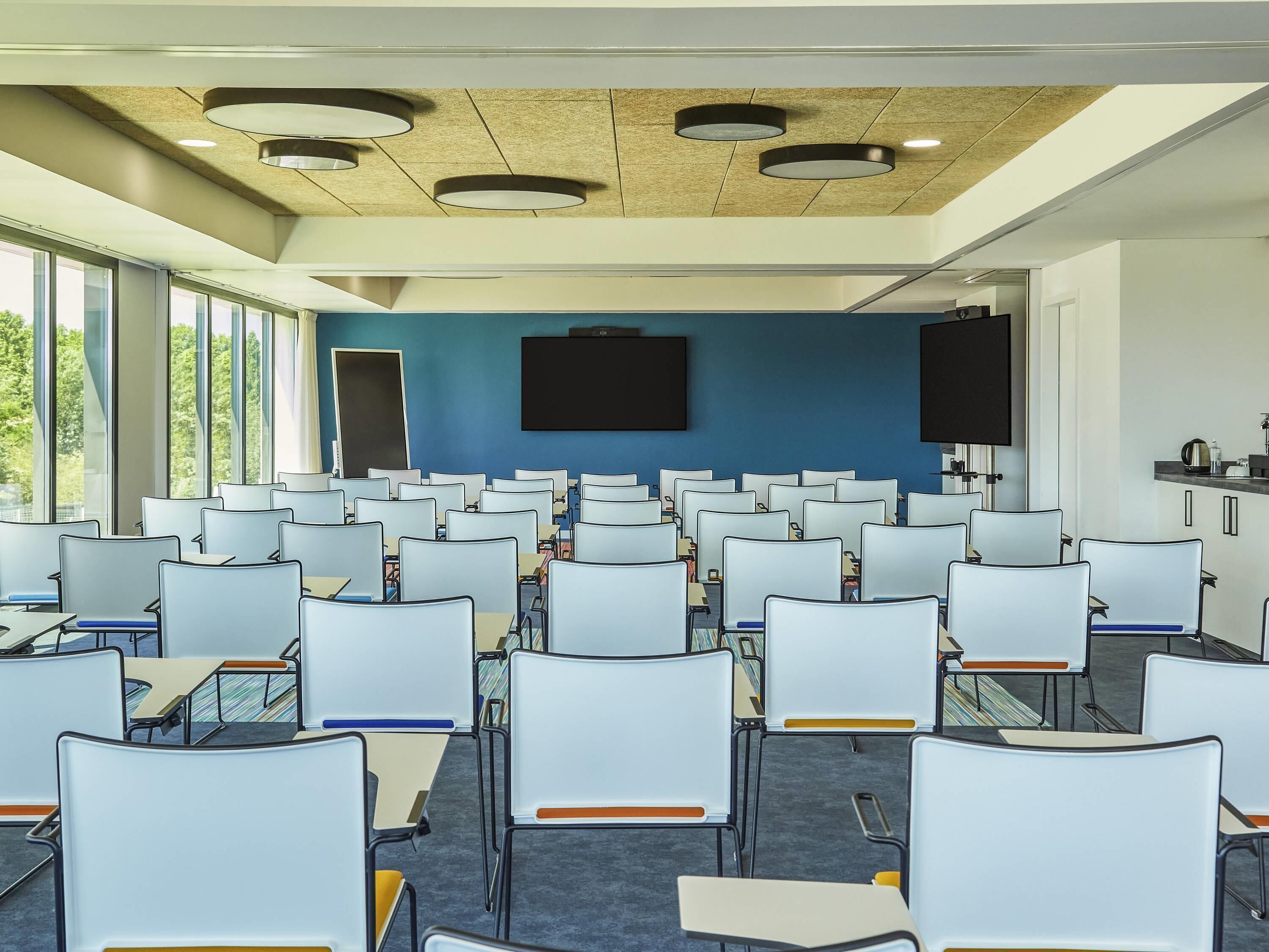 Uma sala de conferência com fileiras de cadeiras brancas, uma tela grande e teto de madeira.