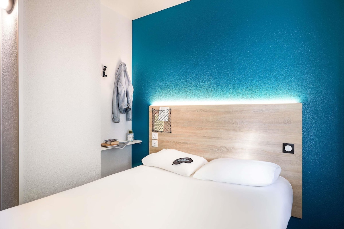Quarto de hotel com cama, mesa de cabeceira, luminária na parede, estante com livros e planta, e uma parede azul decorativa.