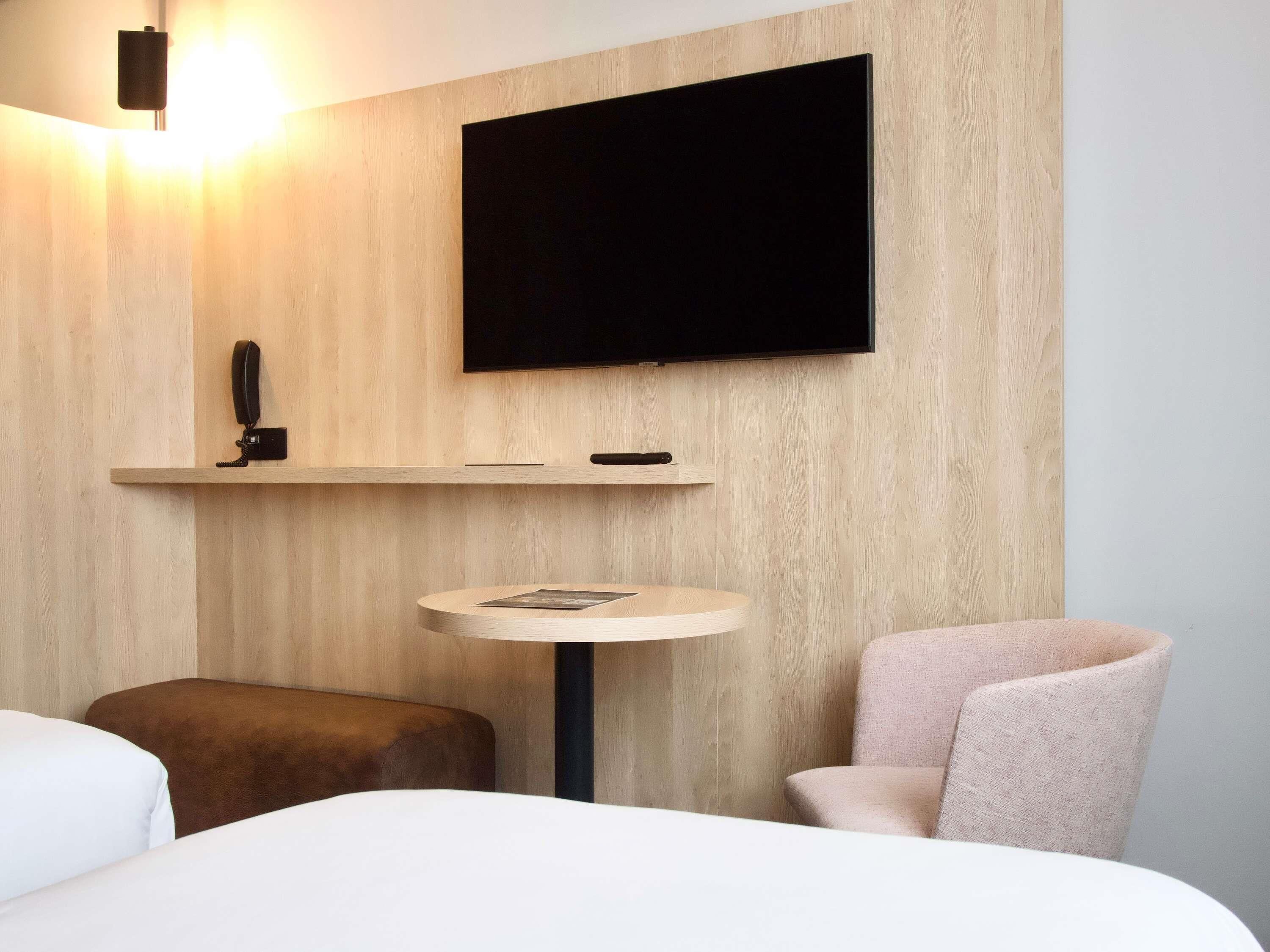 A hotel room with a bed, a round table, a chair, a mounted TV, and a telephone on a shelf.