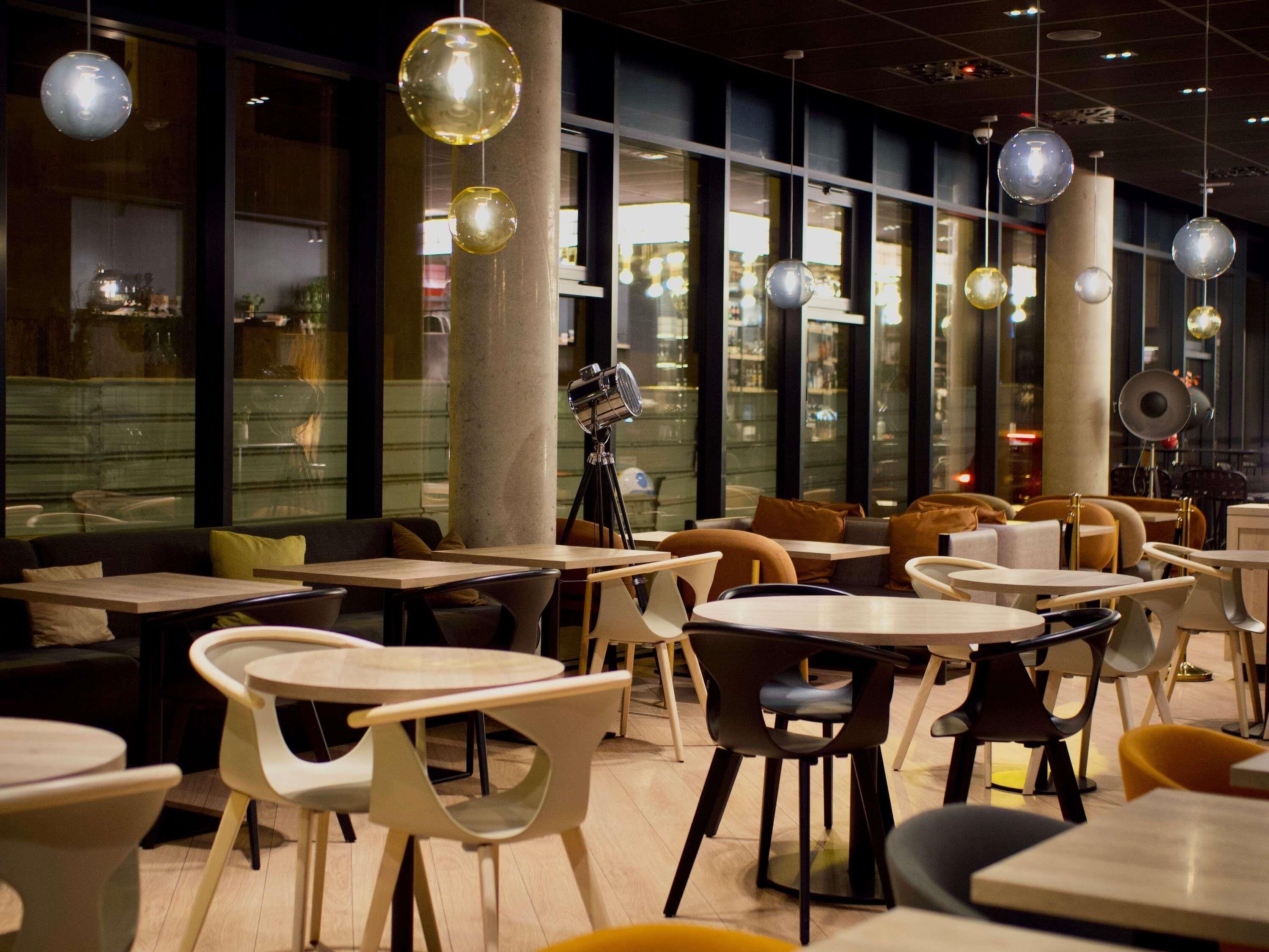 Een moderne eetruimte met ronde tafels en stoelen, grote ramen en hangende sferische lampen.