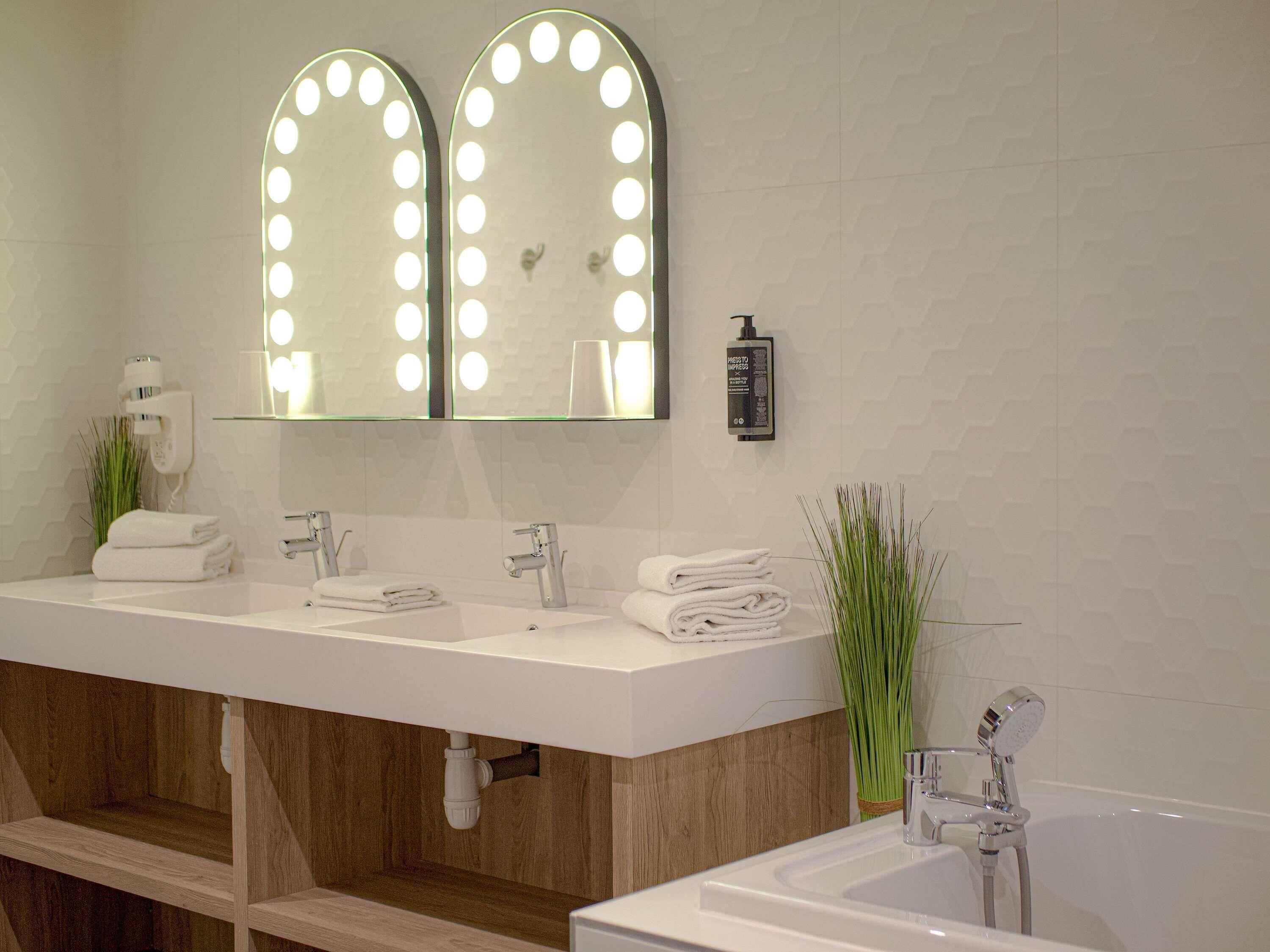 A modern bathroom with two sinks, vanity mirrors with lights, a towel rack, a handheld showerhead, and a toilet paper holder.