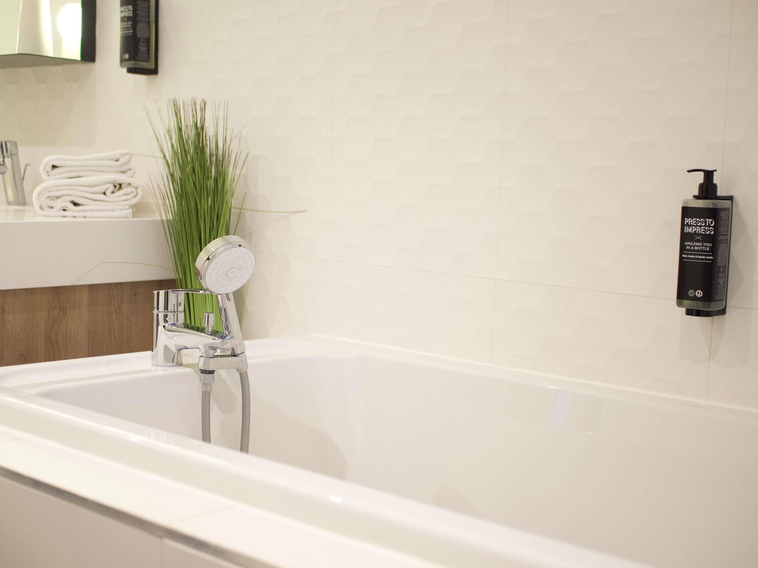 A modern bathroom with a bathtub, a towel rack with folded towels, a green plant, and a wall-mounted bottle dispenser.