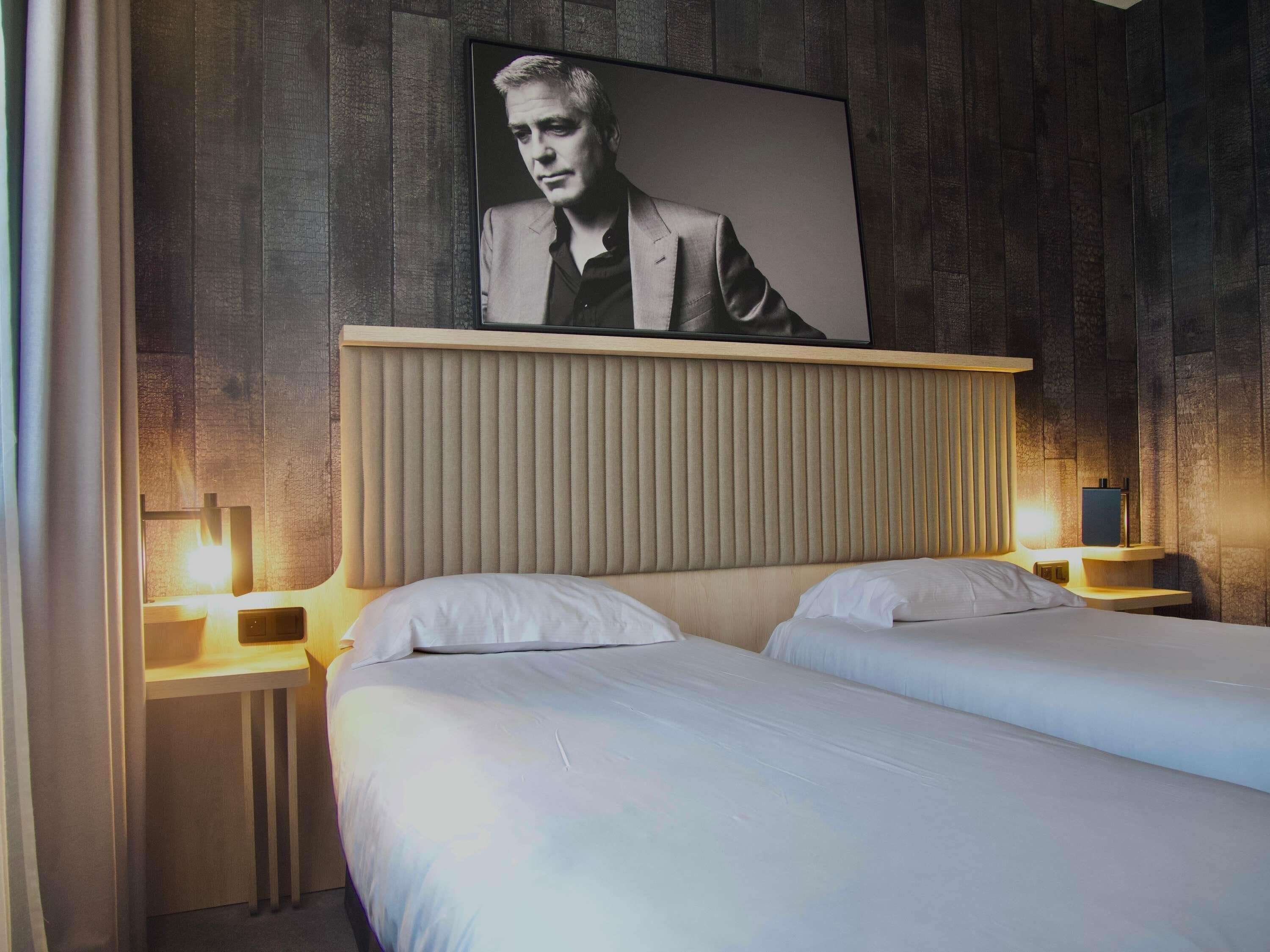 A hotel room with a large bed, two bedside tables, a headboard with vertical slats, and a framed portrait on the wall.