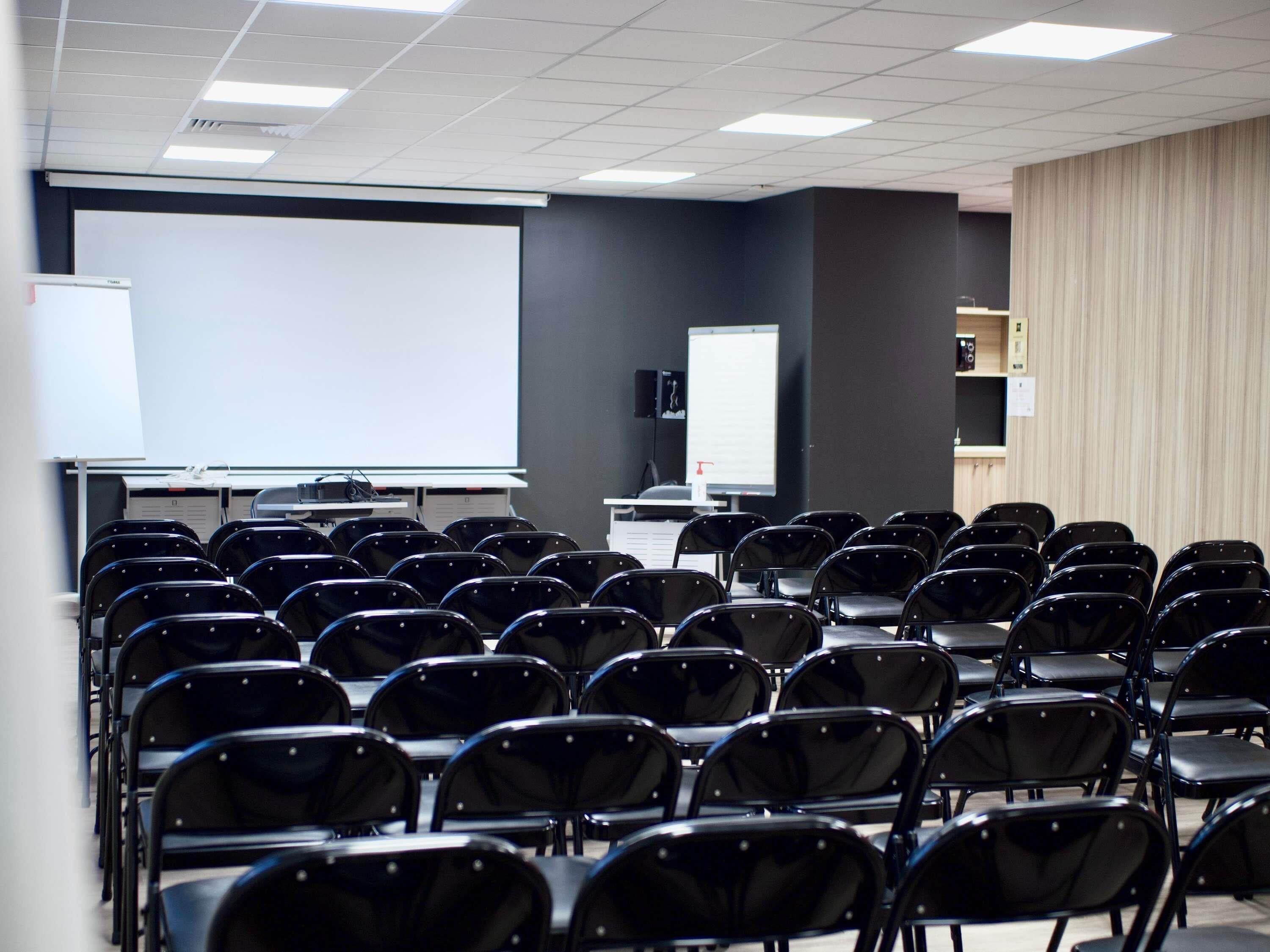 Une salle de conférence avec des rangées de chaises, un écran de projection et un tableau blanc.