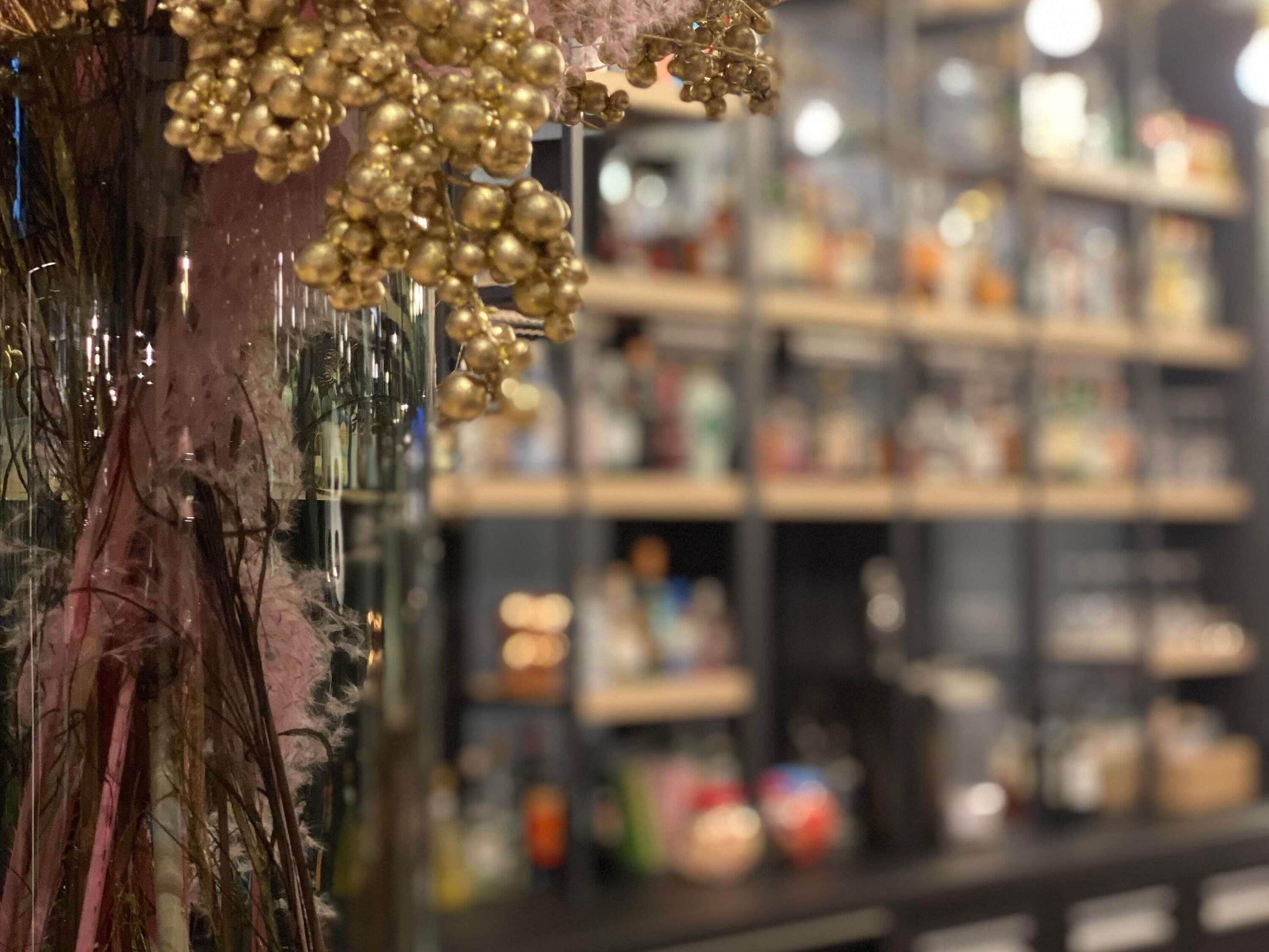 Gros plan d’un vase décoratif en verre, orné de sphères dorées et contenant une plante aux teintes rosées.